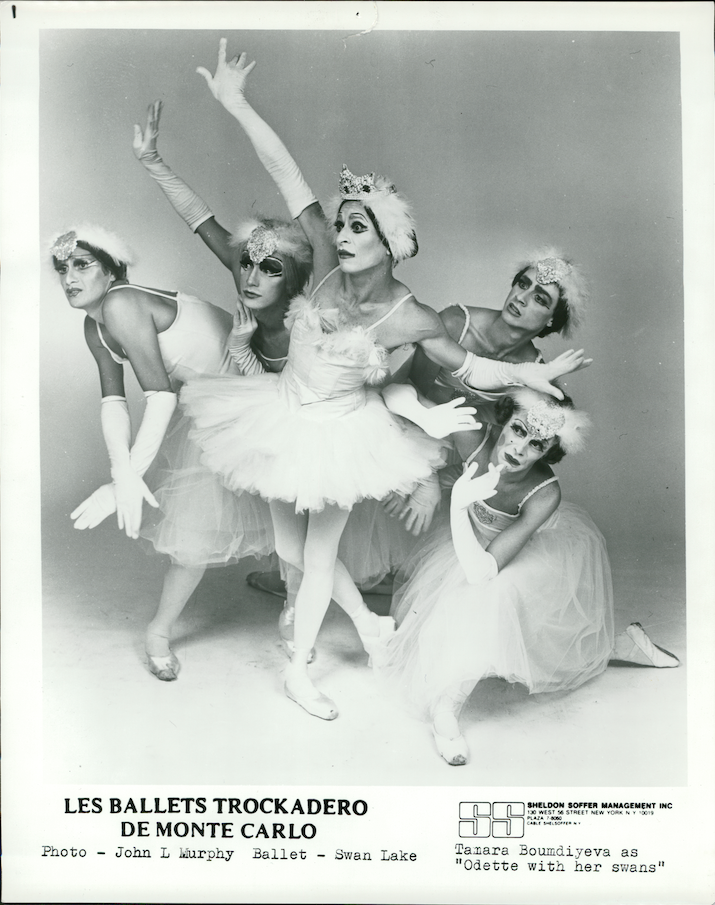 A black and white image of five dancers in drag posing for photo in "Swan Lake." They crowd together, looking fearfully toward the left as the center swan reaches her arms out as if to protect the others. They wear white tutus and feather headpieces.